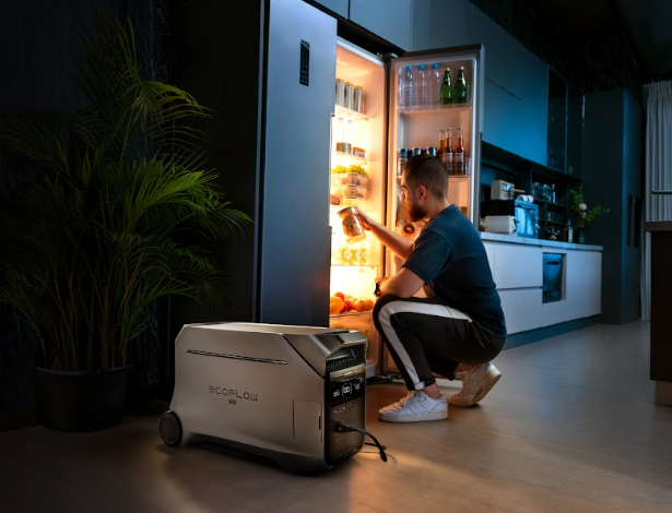 Person sitting on the floor next to an open refrigerator with a portable power station in the foreground.
