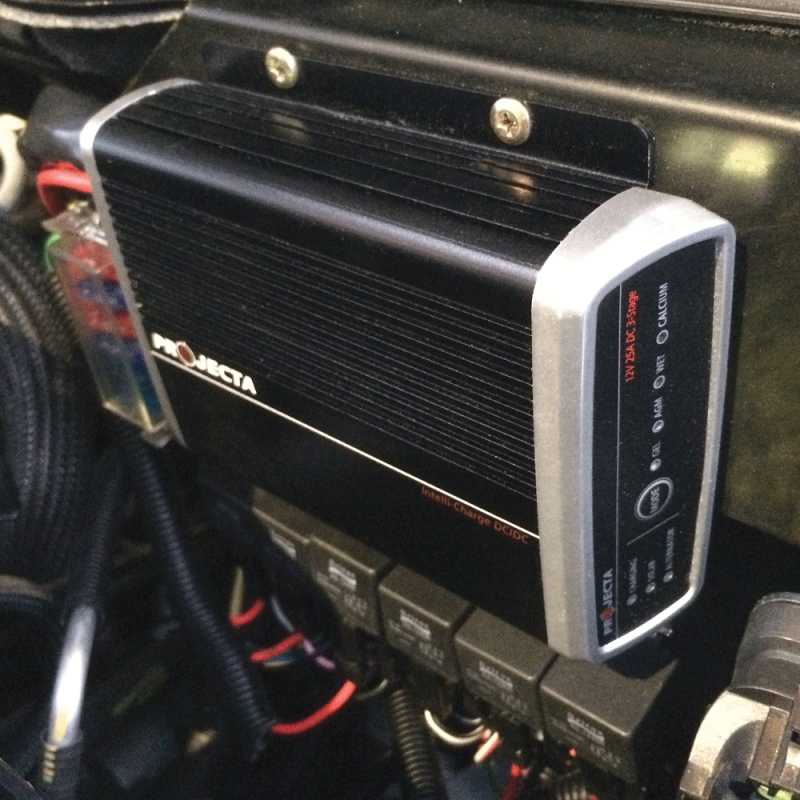 A black Projecta Intelli-Charge DC-DC charger, labeled "12V 25A DC 3-Stage," mounted in an engine bay. It has silver end caps and indicator lights for various battery types (AGM, WET, CALCIUM) and charging status. Wires and what appear to be relays are visible below and around the charger.