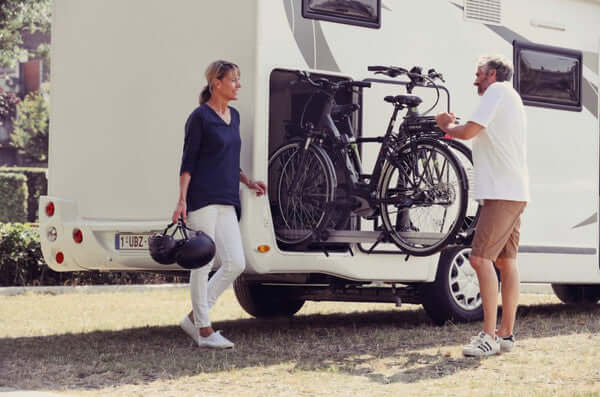 Two people with an RV and bicycles, one person holding helmets.
