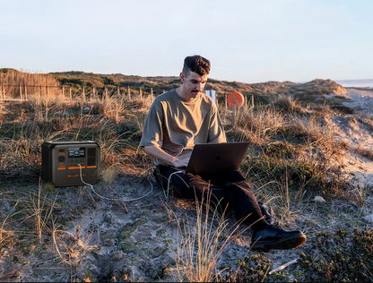 Person sitting outdoors on a sandy dune with a laptop and power station. 