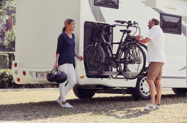 Two people with an RV and bicycles, one person holding helmets.