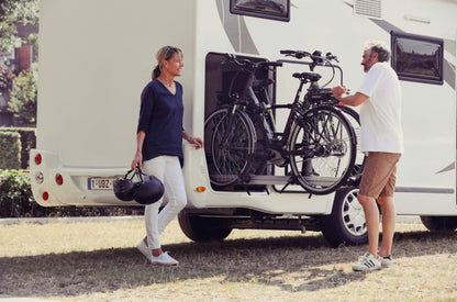 Two people with an RV and bicycles, one person holding helmets.